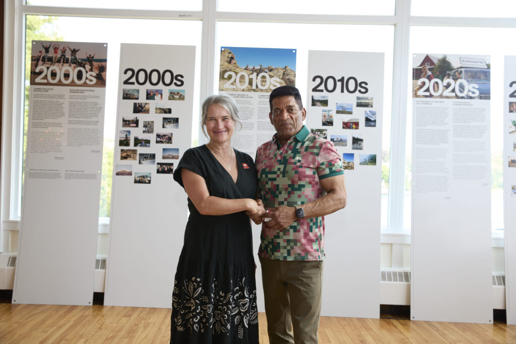 Two people shake hands in front of Katimavik decade-timeline exhibition panels.