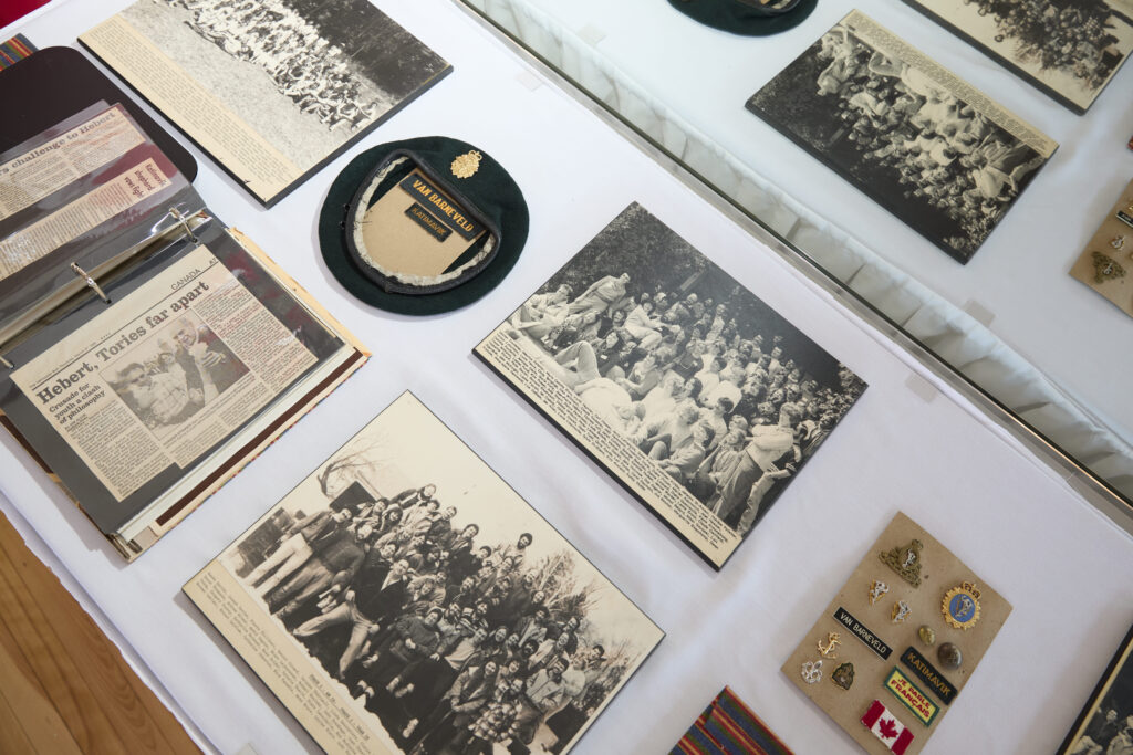 Close-up display of Katimavik historical artifacts including vintage black and white photographs, newspaper clippings, a black beret with the Katimavik patch, and commemorative pins and badges mounted on a white exhibition surface.