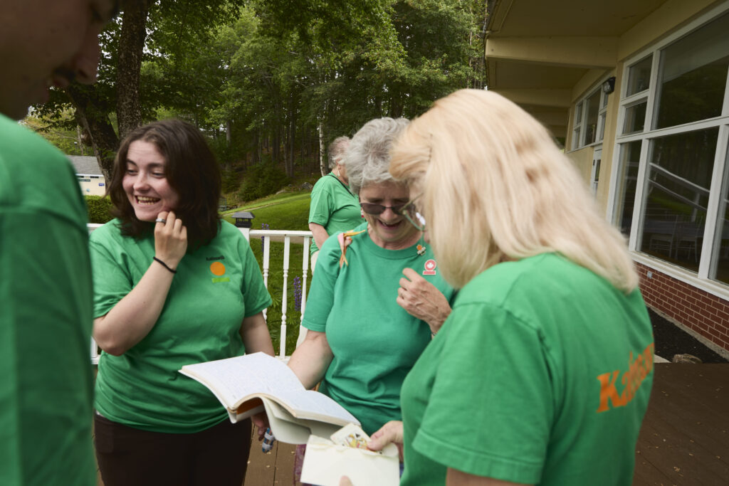 Katimavik participants of various ages in matching green shirts smile while looking at materials together on a residential deck overlooking the forest.